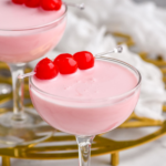 two coupe glasses of Pink Squirrel cocktail garnished with maraschino cherries