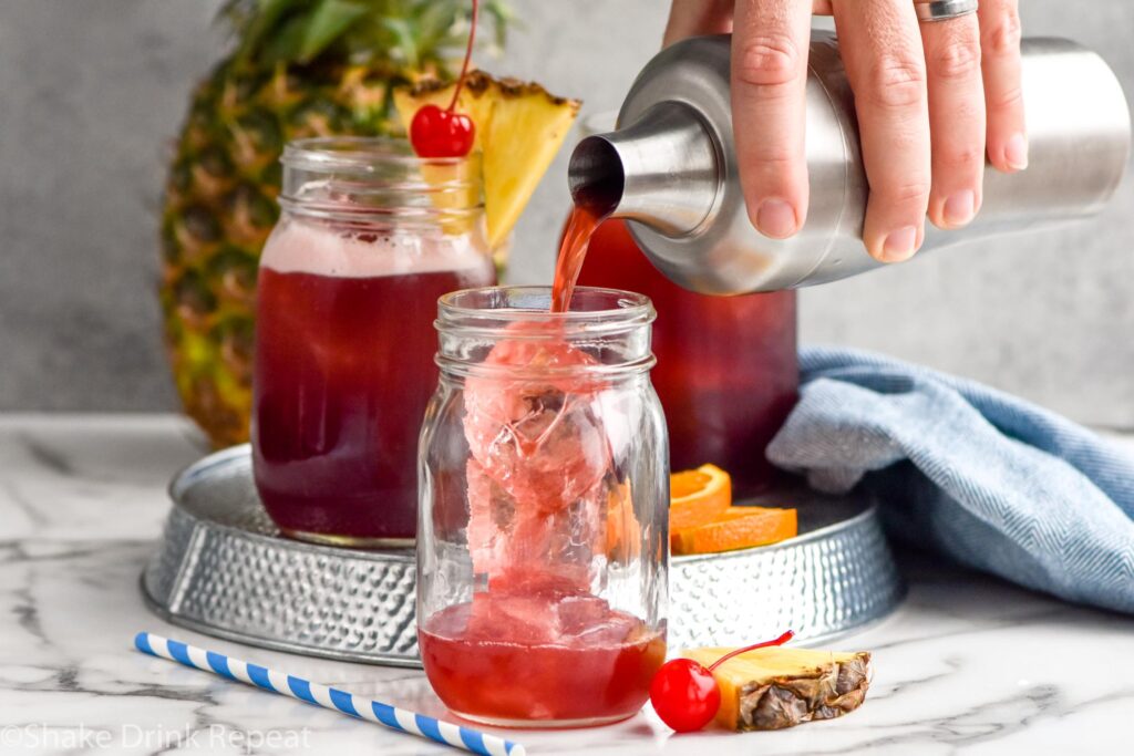 man's hand pouring cocktail shaker of Rum Runner ingredients into a glass with ice surrounded by fresh pineapple, oranges, and a cherry