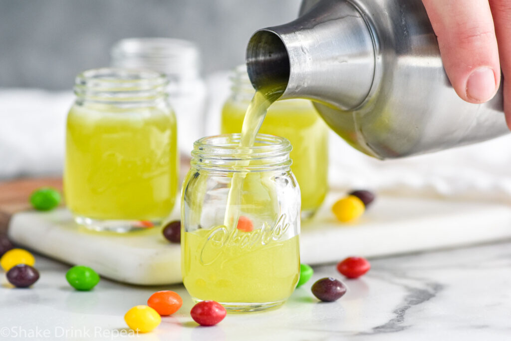 man's hand pouring cocktail shaker of Skittles Shot ingredients into a shot glass surrounded by skittles candy.