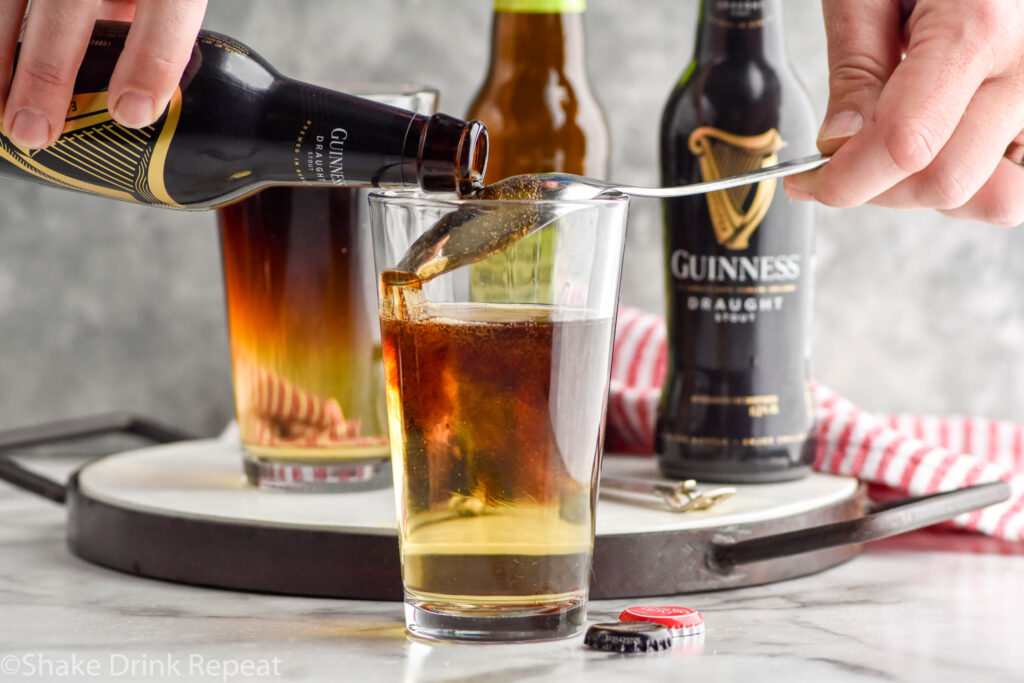 man's hands pouring bottle of Guinness over a spoon into a glass of hard cider to make a Snakebite drink