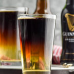 two pint glasses of Snakebite drink with bottles of hard cider and Guinness in the background