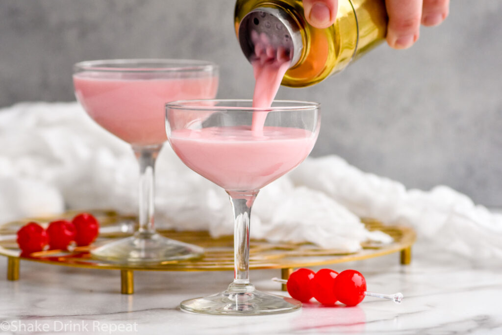 cocktail shaker of Pink Squirrel ingredients pouring into a coupe glass surrounded by cherries