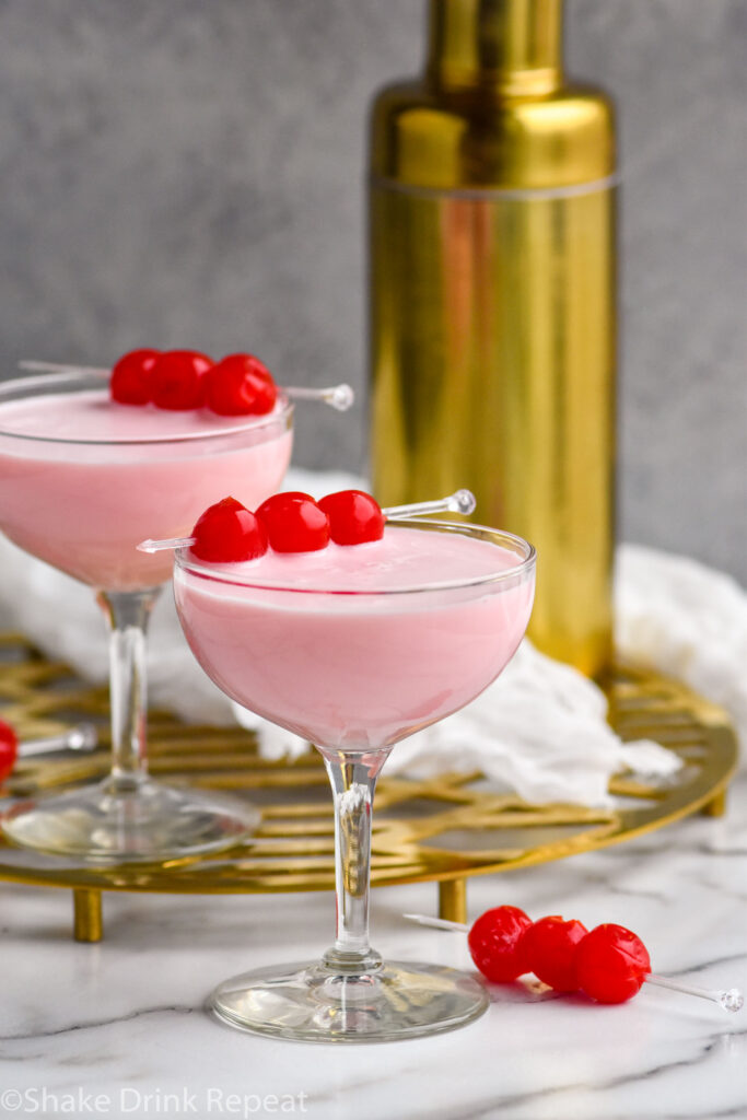 Two coupe glasses of Pink Squirrel cocktail garnished with maraschino cherries, gold cocktail shaker in the background