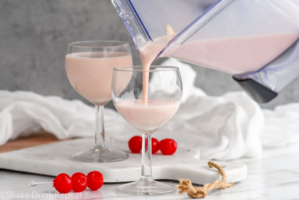 blender pouring Pink Squirrel with Ice Cream ingredients into a glass surrounded by maraschino cherries