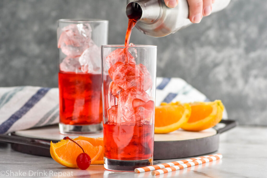 man's hand pouring a cocktail shaker of Alabama Slammer ingredients into a glass of ice surrounded by orange slices, a cherry, and two straws