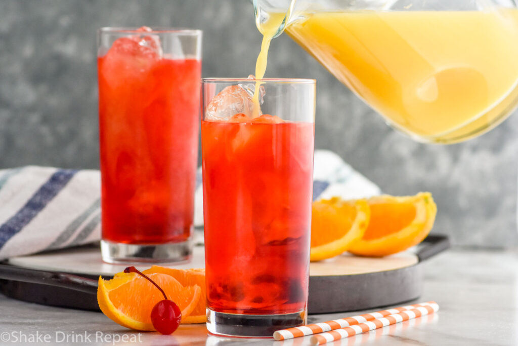 glass pitcher of orange juice pouring into a glass of Alabama Slammer ingredients with ice, orange slices, cherry, and straws in the background