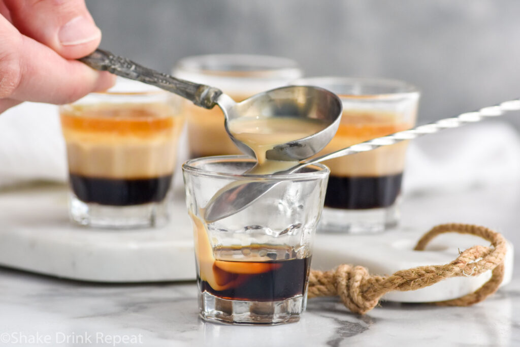 man's hand pouring Baileys over the back of a spoon into a shot glass of Kahlua to create a Duck Fart Shot