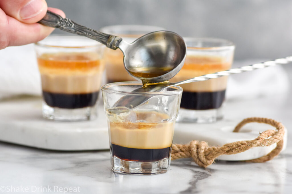 Man's hand pouring whiskey over the back of a spoon into a shot glass over Kahlua and Baileys to make a Duck Fart Shot