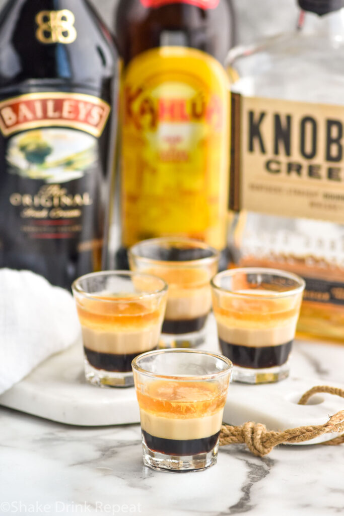 four shot glasses of Duck Fart shot recipe with bottles of whiskey, Baileys, and Kahlua in the background