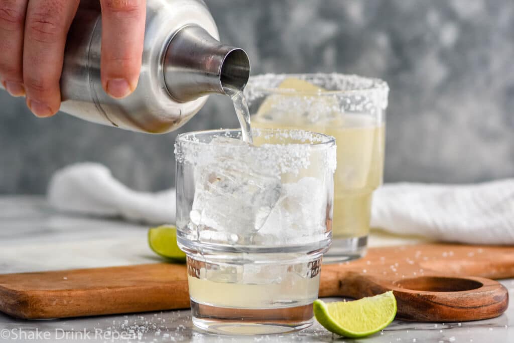 Man's hand pouring golden margarita from shaker bottle into clear glass with ice and a salted rim. Lime wedges lay beside the class on the counter for garnish.