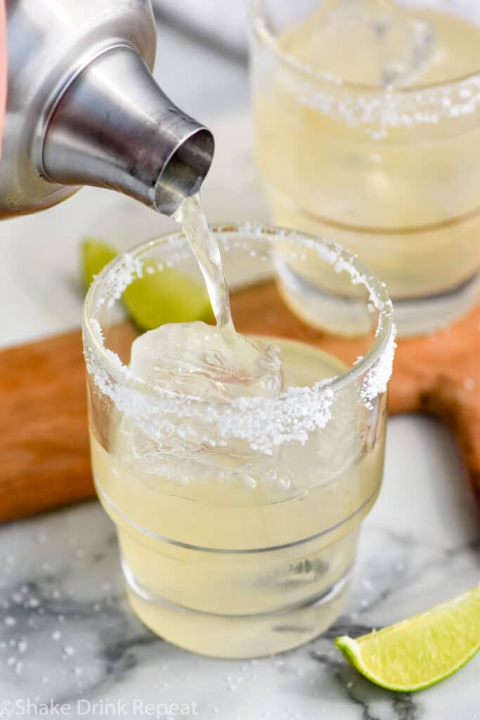 Golden margarita being poured from shaker bottle into clear glass with ice and a salted rim. Lime wedges lay beside the class on the counter for garnish.