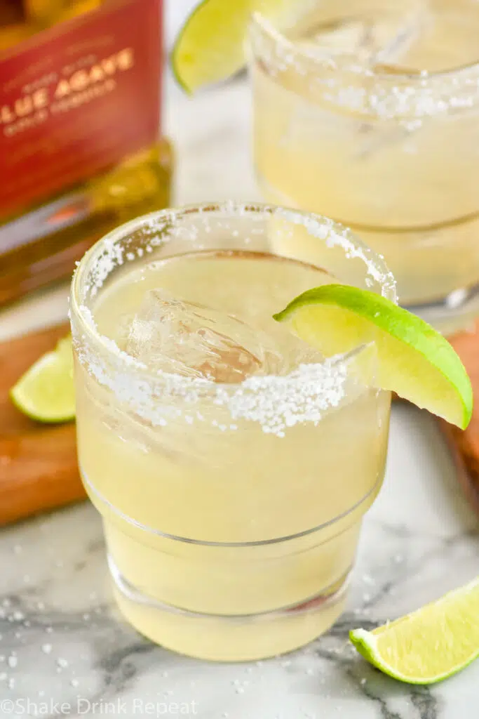 Golden margarita served in a clear glass over ice with a salted rim and lime wedge garnish. Bottle of gold tequila sits in the background.