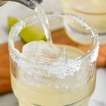 Pinterest graphic of golden margarita recipe. Text says, "Tequila, Golden Margarita, and shakedrinkrepeat.com" Photo is of silver shaker pouring golden margarita into a clear glass with ice and a salted rim. Lime wedges for garnish lay on the counter beside the glass.