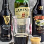pint glass of Guinness next to a shot glass of whiskey and baileys to make an irish car bomb recipe. Bottles of Guinness, Jameson, and Baileys in the background