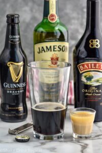 pint glass of Guinness next to a shot glass of whiskey and baileys to make an irish car bomb recipe. Bottles of Guinness, Jameson, and Baileys in the background