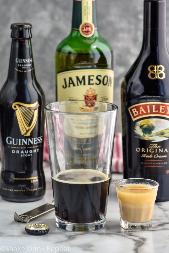 pint glass of Guinness next to a shot glass of whiskey and baileys to make an irish car bomb recipe. Bottles of Guinness, Jameson, and Baileys in the background