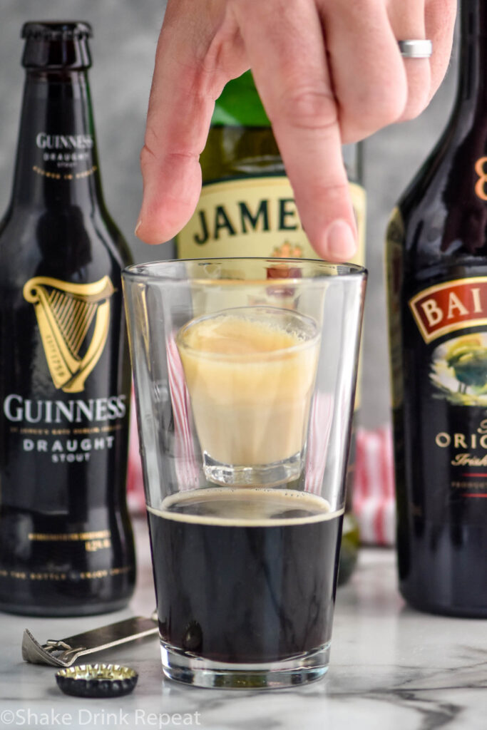 man's hand dropping a shot glass of whiskey and baileys into a glass of Guinness to make an Irish car bomb recipe, bottles of Guinness, Jameson, and Baileys sit in the background