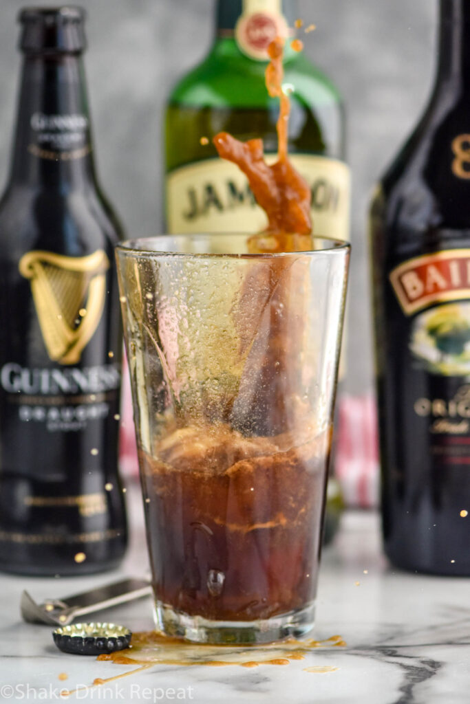 shot glass of whiskey and baileys dropping into a glass of Guinness to make an Irish car bomb recipe, bottles of Guinness, Jameson, and Baileys sit in the background