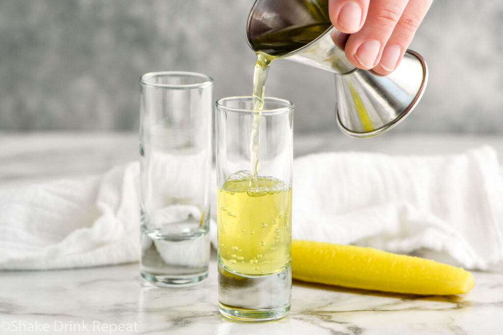 man's hand pouring cocktail jigger of pickle juice into a shot glass to make a pickleback