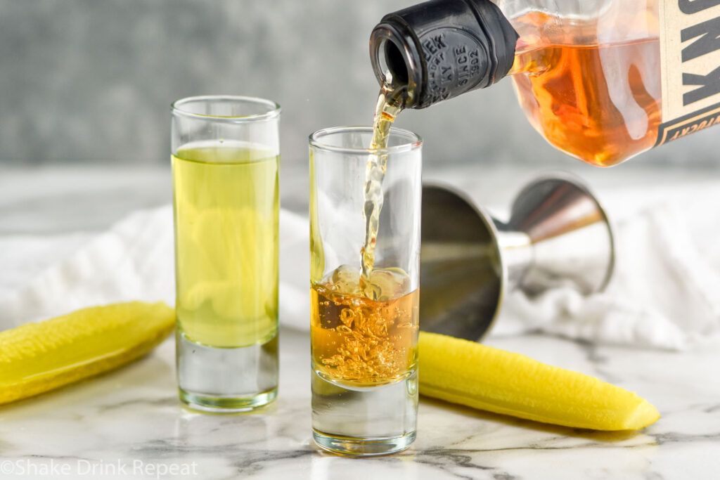 bottle of whiskey pouring into a shot glass next to a shot glass of pickle brine to make a pickleback with two pickles next to glasses