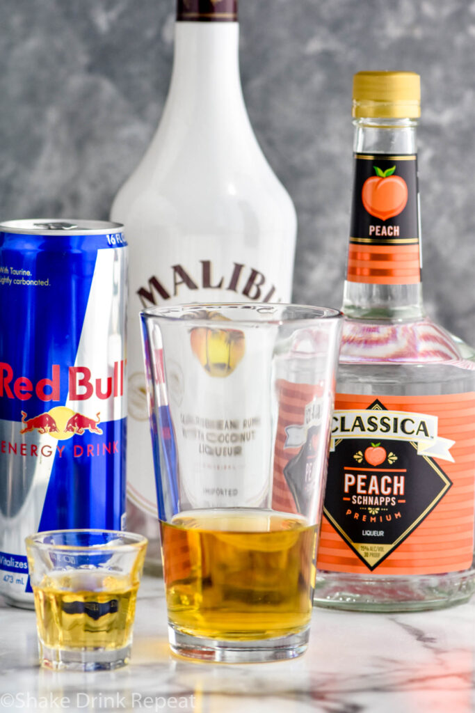 shot glass of liquor next to a glass of red bull to make a vegas bomb shot with bottles of Malibu rum, peach schnapps, land red bull in the background
