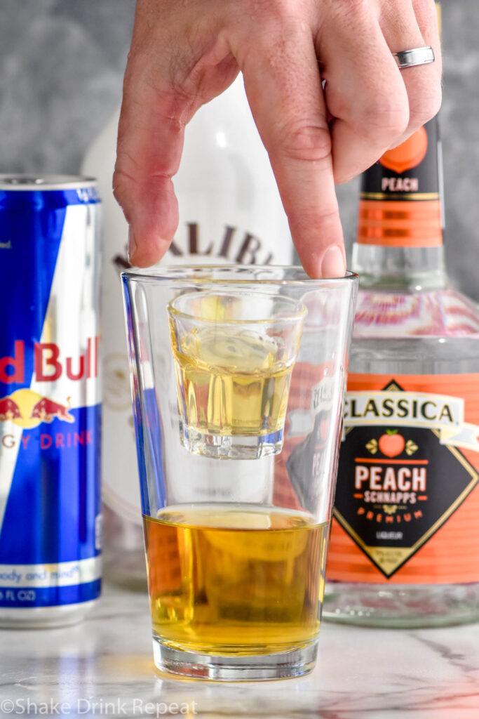 man's hand dropping a shot glass of liquor into a glass of red bull to create a vegas bomb shot with bottles of peach schnapps, malibu and red bull in the background