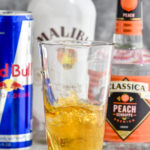 shot glass of liquor dropped into a glass of red bull to make a Vegas Bomb shot with bottles of peach schnapps, malibu and red bull in the background