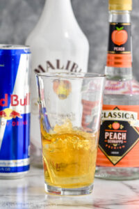 shot glass of liquor dropped into a glass of red bull to make a Vegas Bomb shot with bottles of peach schnapps, malibu and red bull in the background