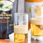 Pinterest graphic of buttery nipple shot recipe. Text says, "Buttery Nipple shakedrinkrepeat.com" Image is of two shot glasses of buttery nipple shot recipe. Bottles of Irish cream and butterscotch schnapps in the background.