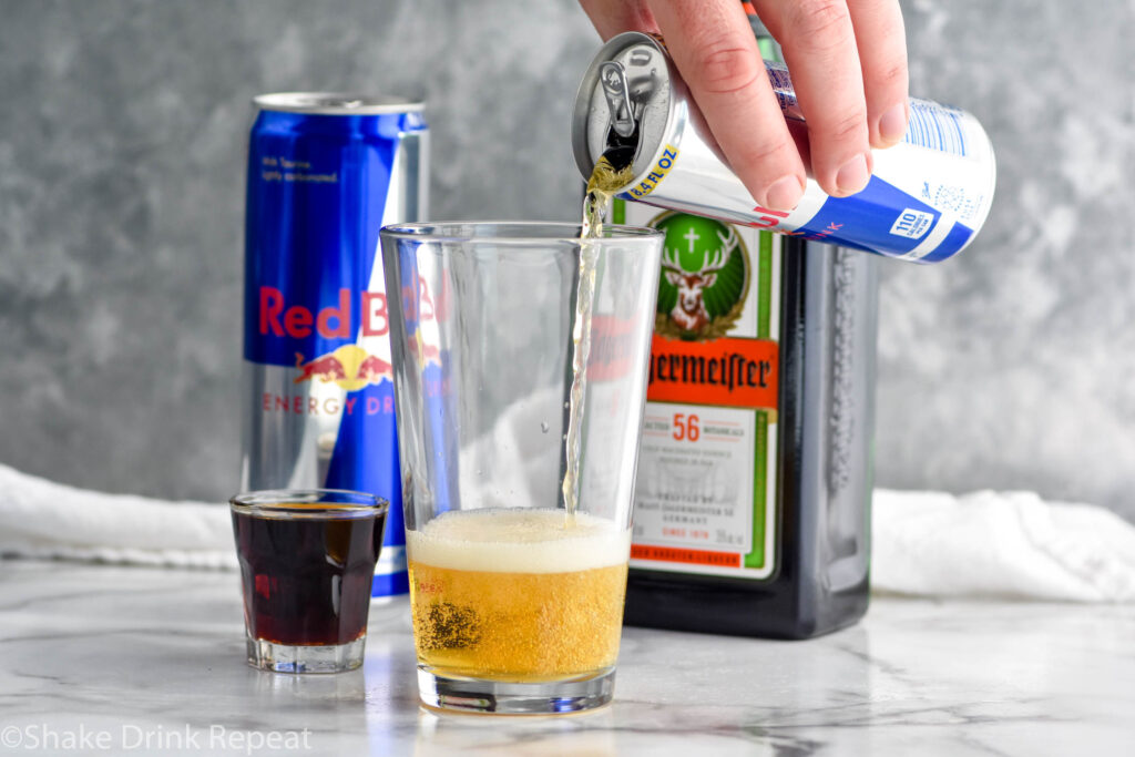 man's hand pouring can of Red Bull energy drink into a pint glass with shot glass of Jägermeister sitting beside. Can of Red bull and bottle of Jägermeister sit in background