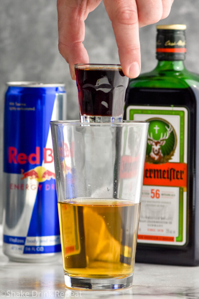 Man's hand holding shot glass of Jägermeister over pint glass of Red Bull to make a Jager Bomb shot, bottle of Jägermeister and can of Red Bull sit in background