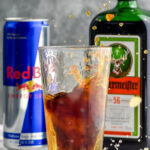 shot glass of Jägermeister splashing into pint glass of Red Bull to make a Jager Bomb, bottle of Jägermeister and can of Red Bull sit in background
