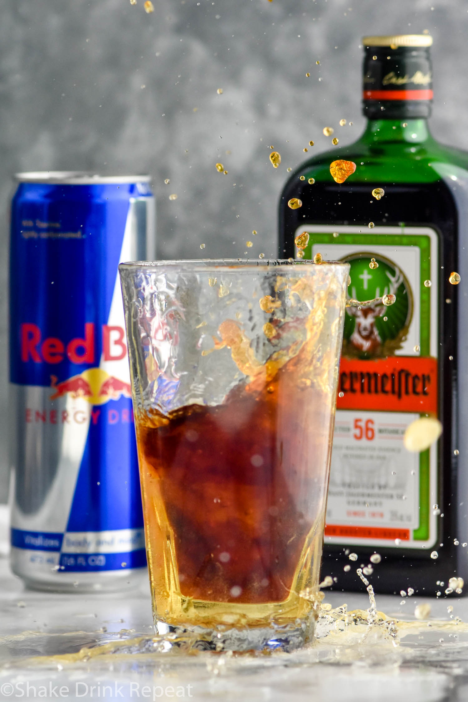 shot glass of Jägermeister splashing into pint glass of Red Bull to make a Jager Bomb, bottle of Jägermeister and can of Red Bull sit in background