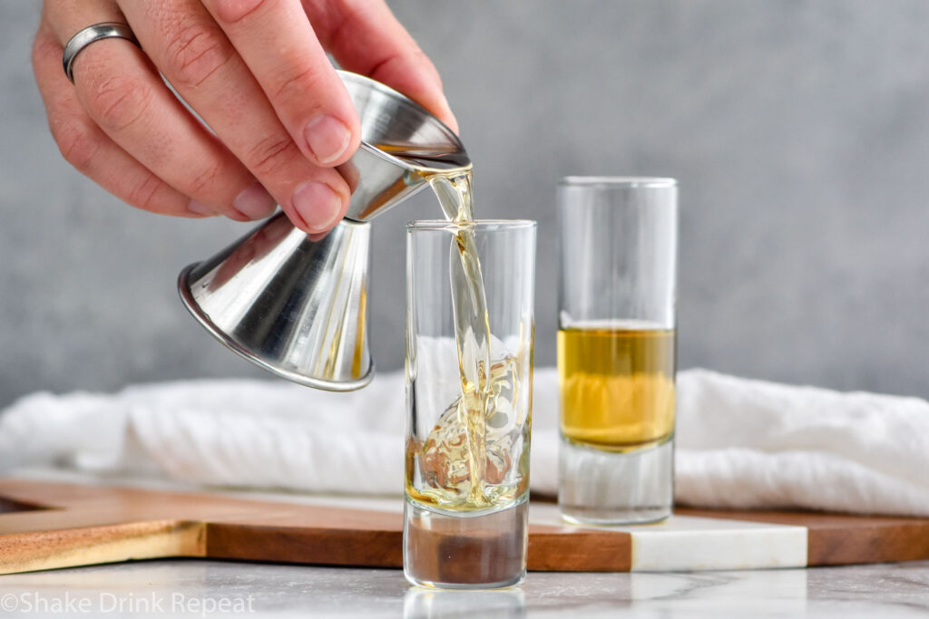 Photo of man's hand pouring butterscotch schnapps into a shot glass for buttery nipple recipe. Second shot glass with butterscotch schnapps sits on the counter.