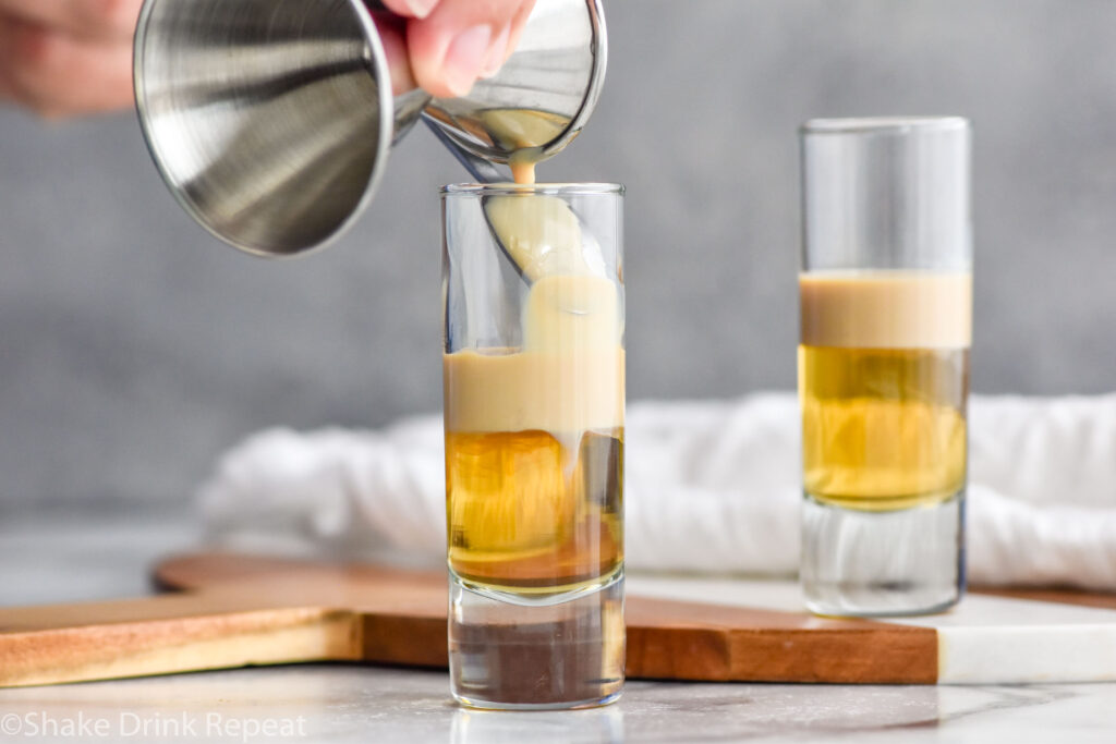 Photo of man's hand pouring Irish cream over a spoon into a shot glass with butterscotch schnapps for buttery nipple recipe. Second shot glass with buttery nipple shot is in the background.