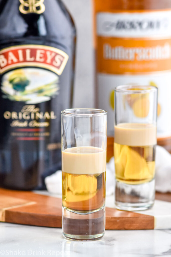 Photo of two shot glasses of buttery nipple shot recipe with a bottle of Irish cream and of butterscotch schnapps in the background.