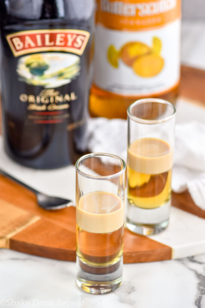 Overhead photo of two shot glasses of Buttery Nipple Shot recipe with a bottle of Irish cream and of butterscotch schnapps in the background.