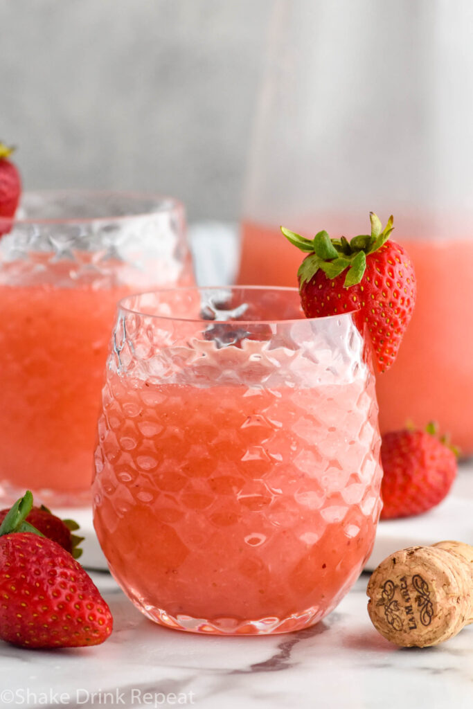 glass of Frose cocktail garnished with and surrounded by fresh strawberries, pitcher and glass of Frose sit in background