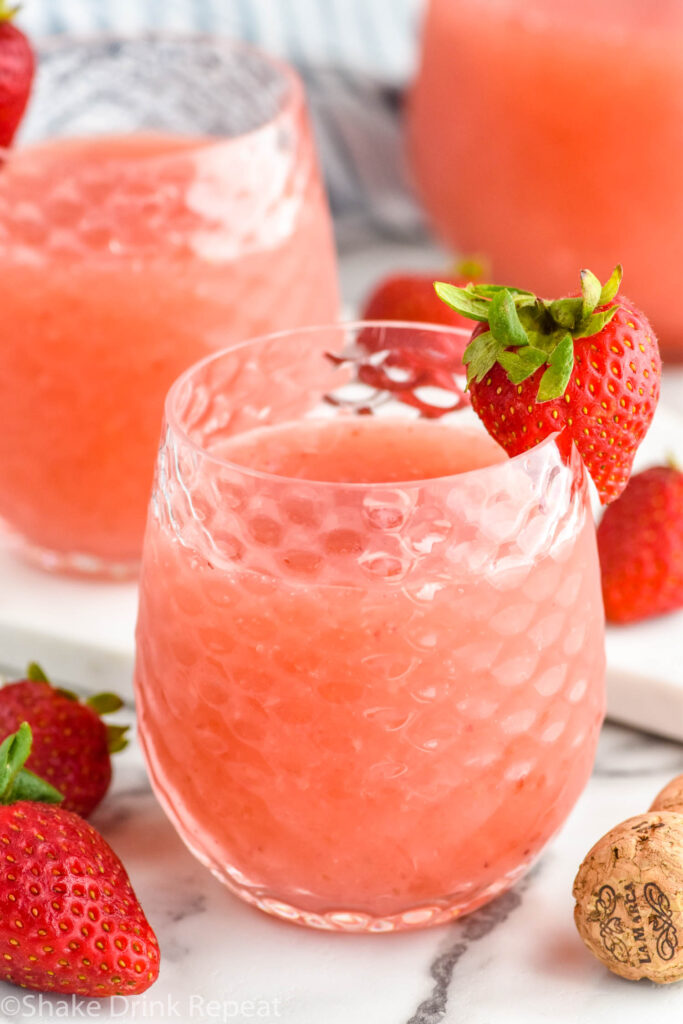 glass of Frose cocktail garnished with and surrounded by fresh strawberries, pitcher and glass of Frose sit in background