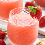 glass of Frose cocktail garnished with and surrounded by fresh strawberries, pitcher and glass of Frose sit in background