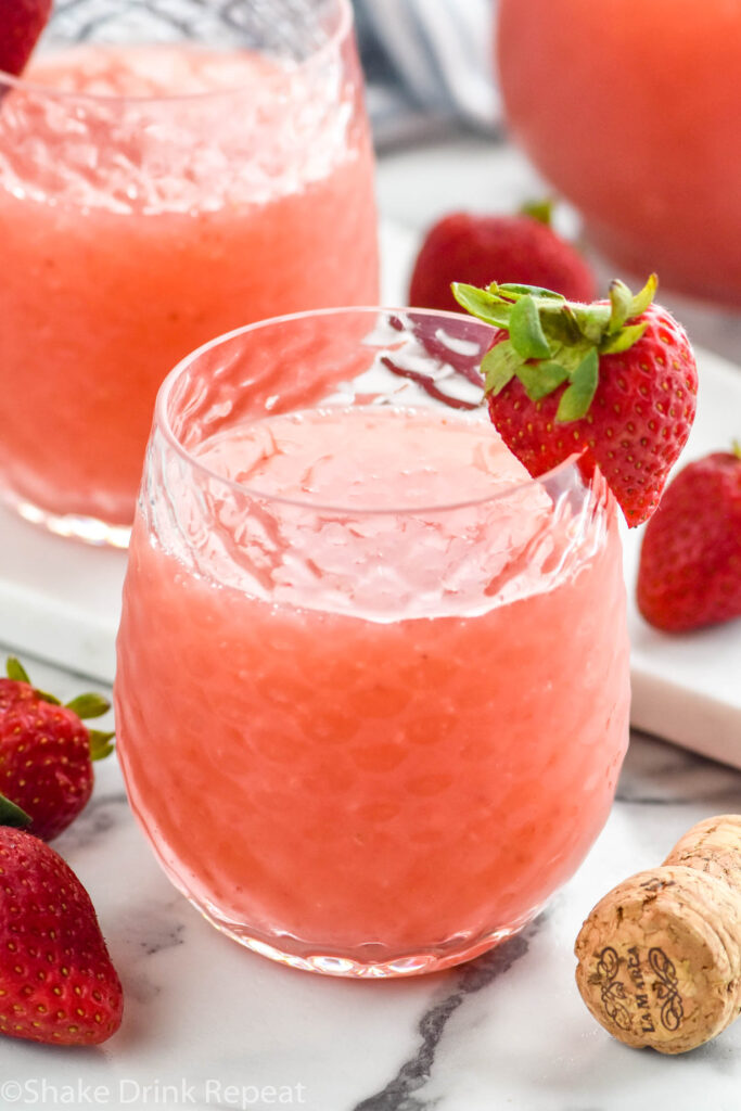 glass of Frose cocktail garnished with and surrounded by fresh strawberries, pitcher and glass of Frose sit in background