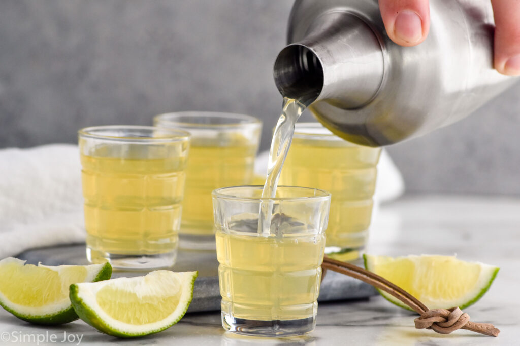 man's hand pouring cocktail shaker of green tea shot ingredients into a shot glass surrounded by other green tea shots and lime wedges