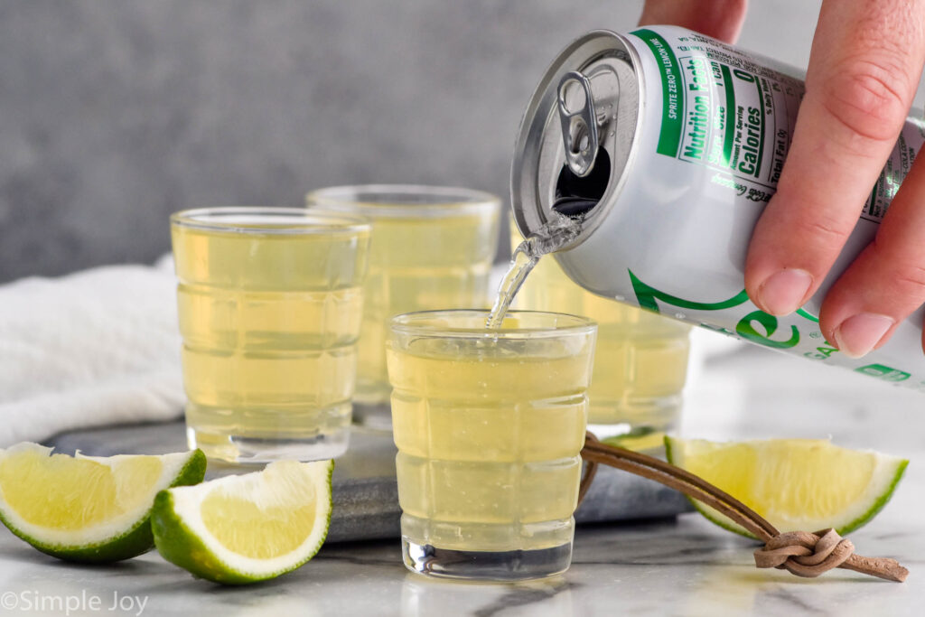 man's hand pouring can of Sprite into a shot glass of green tea shot ingredients surrounded by other green tea shots and lime wedges