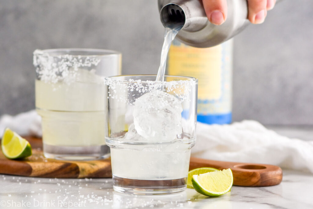 Photo of man's hand pouring Mezcal Margarita from a shaker into a glass with ice and a salted rim. Lime wedges and a bottle of mezcal sit on the counter in the background.