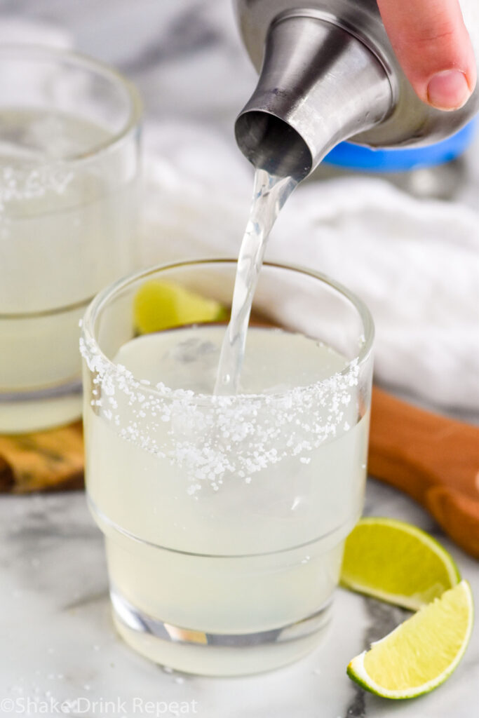 Photo of man's hand pouring Mezcal Margarita from a shaker into a glass with ice and a salted rim. Lime wedges sit on the counter beside the glass.