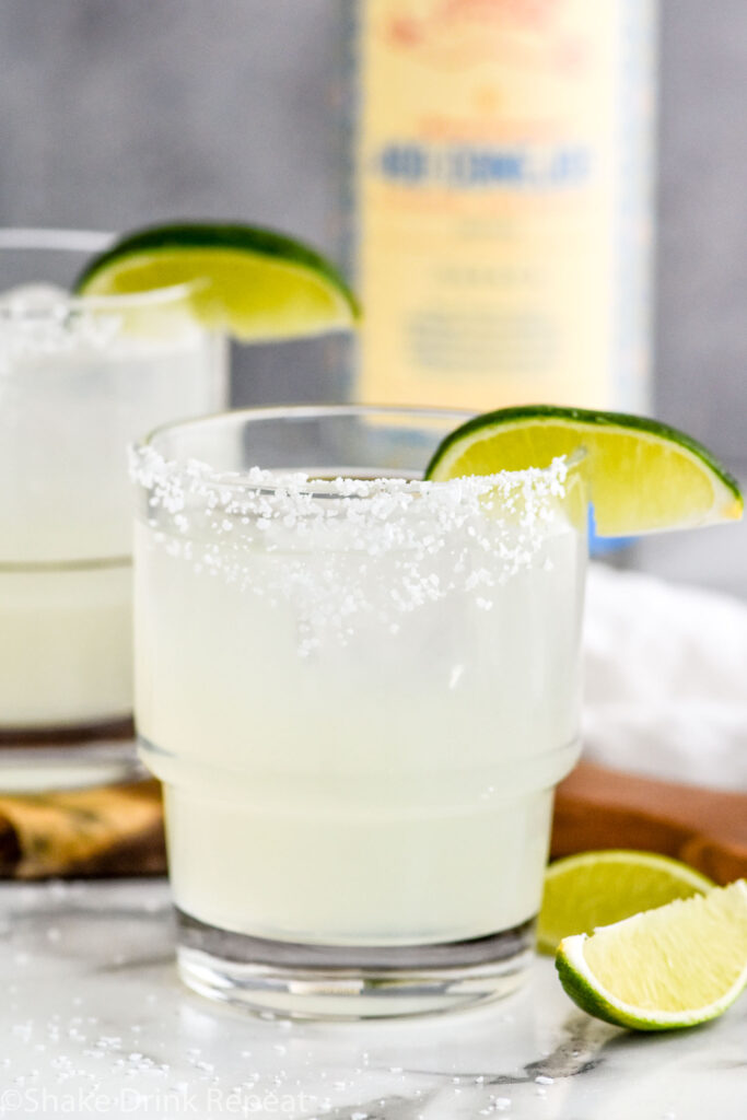 Photo of two glasses of Mezcal Margaritas garnished with salted rims and lime wedges. A bottle of mezcal sits on the counter in the background.