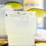 Photo of two glasses of Mezcal Margaritas garnished with salted rims and lime wedges. A bottle of mezcal sits on the counter in the background.