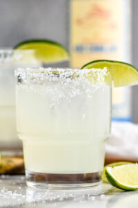 Photo of two glasses of Mezcal Margaritas garnished with salted rims and lime wedges. A bottle of mezcal sits on the counter in the background.