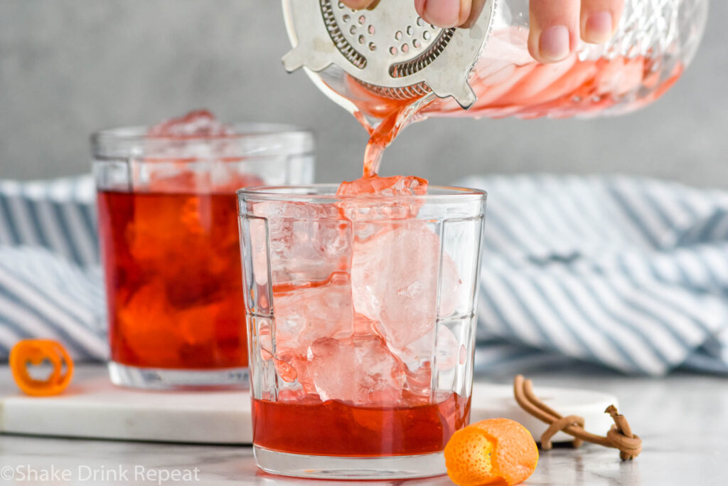 Photo of man's hand pouring mezcal negroni through a strainer into a glass tumbler with ice. Orange peels lay beside the glass for garnish.