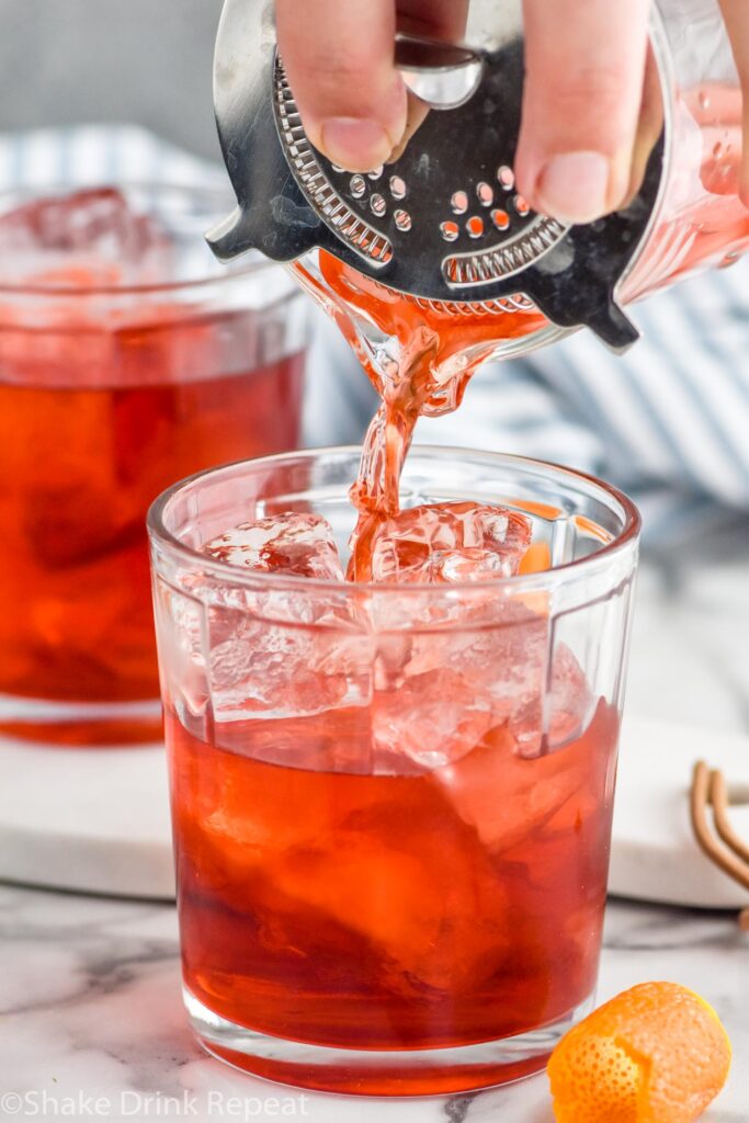 Photo of man's hand pouring mezcal negroni through a strainer into a glass tumbler with ice. Orange peels lay beside the glass for garnish.